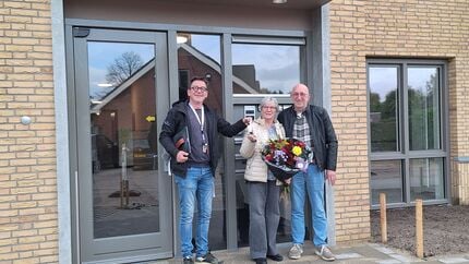 Drie mensen staan buiten voor een modern gebouw met lichtkleurige bakstenen en grote ramen. Eén persoon houdt een kleurrijk boeket bloemen vast, wat wijst op een feestelijke gelegenheid. De sfeer is informeel en vriendelijk.