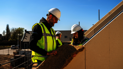 Twee personen met witte bouwhelmen en veiligheidsvesten plaatsen isolatiemateriaal tegen een dakconstructie op een bouwplaats.