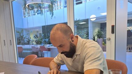 Persoon zit aan een tafel en ondertekent een document in een moderne vergaderruimte met glazen wanden en groene planten op de achtergrond.