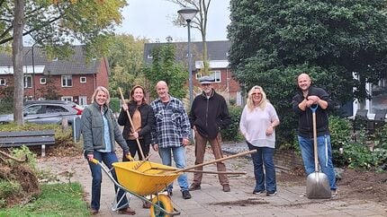 Groep mensen staat buiten op een betegeld pad met tuingereedschap en een gele kruiwagen.